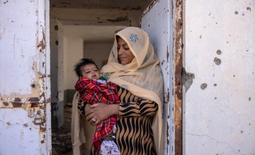 Nasima* (holds her son Rahmin* (2 months old) standing in the doorframe of what was once Sangin's clinic, bombed out and destroyed. There is currently no healthcare available. Photo: Stefanie Glinski