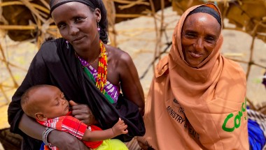 Two women smile at the camera as one of them breastfeeds her baby