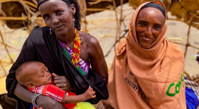 Two women smile at the camera as one of them breastfeeds her baby