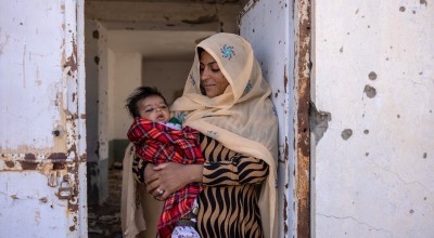 Nasima* (holds her son Rahmin* (2 months old) standing in the doorframe of what was once Sangin's clinic, bombed out and destroyed. There is currently no healthcare available. Photo: Stefanie Glinski